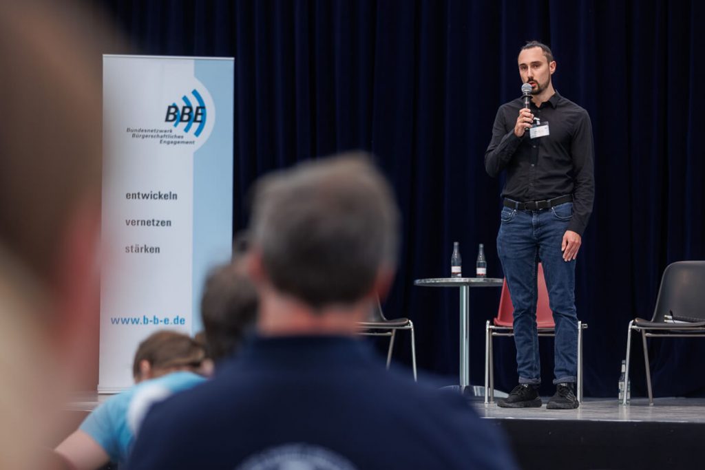 Philipp Hill steht auf einer Bühne und hält eine Rede. Blick aus dem Publikum auf die Bühne.