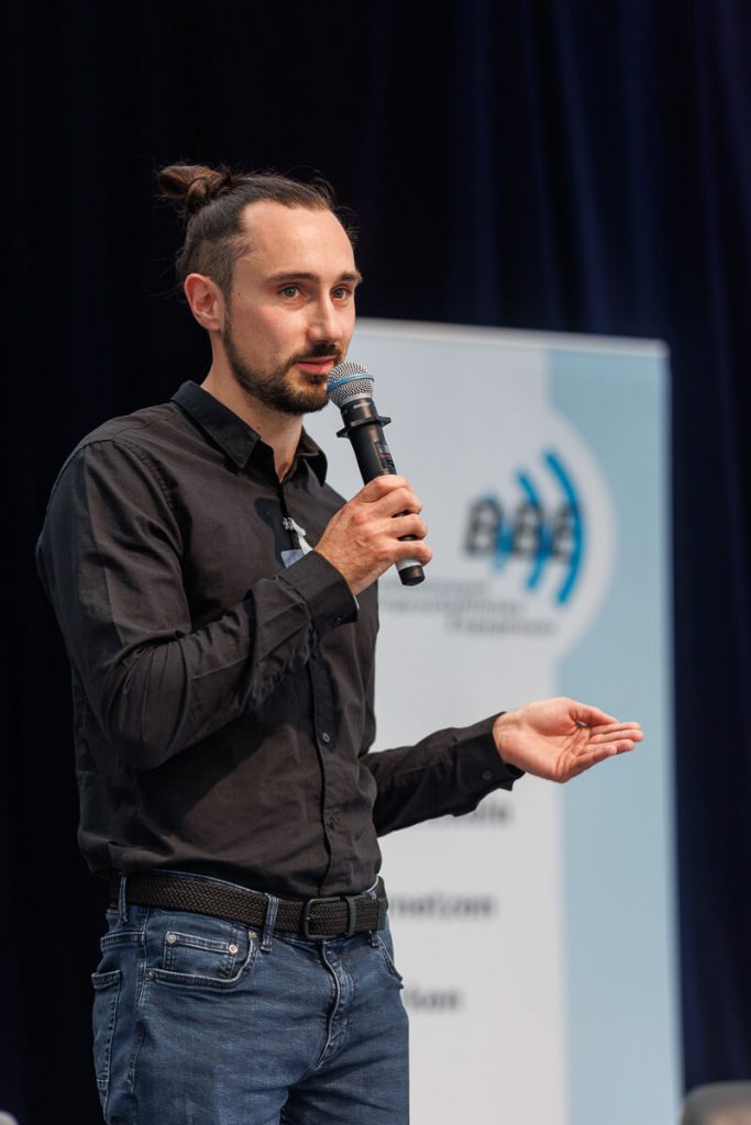 Philipp hält eine Rede. Er hält ein MIkrofon in der Hand. Bühne im Hintergrund ist unscharf.